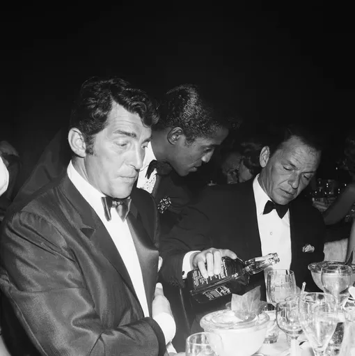 Singer Dean Martin and Sammy Davis, Jr. sit at a table as fellow singer and Rat Pack member Frank Sinatra pours Jack Daniels from a bottle at the Cocoanut Grove during Eddie Fisher's opening night on July 25, 1961 in Los Angeles, California. (Photo by Earl Leaf/Michael Ochs Archives/Getty Images)