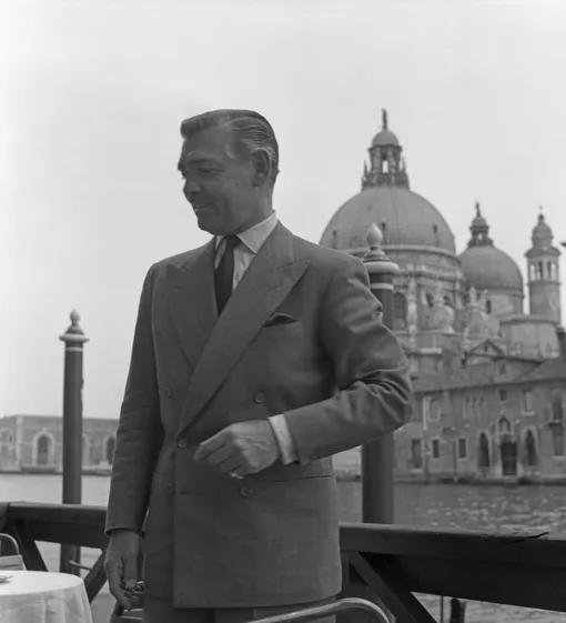 American actor Clark Gable, wearing a suit and a tie, portrayed standing on a restaurant terrace, on the background the Salute Basilica, Venice, 1953. КРЕДИТ s