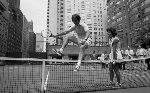 July 11, 1973 Wimbledon whiz Billie Jean King is glad to lend a hand to a doddering 55-year-old-Bobby Riggs-as he hops over net at E. 56th St. court. Bobby and Billie have a date for a $100,000 match one of these days and it's made more interesting in that Billie seems to think Bobby might be some kind of male chauvinist. (Photo by )