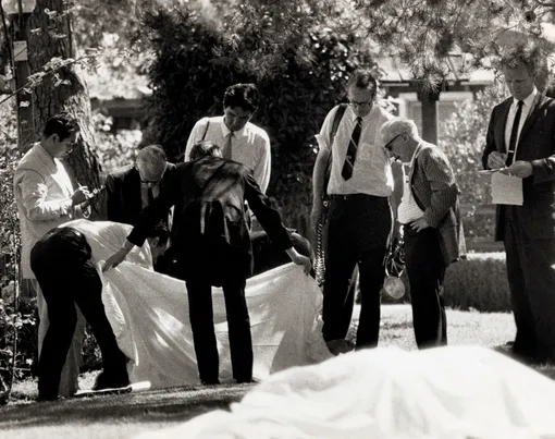 County Coroner Thomas Nogouchi (left) takes notes as assistants hold sheet which covered one body as the other of the two bodies found on the lawn of the Sharon Tate-Roman Polanski home shows in foreground. Five persons were discovered murdered on the property early August 9th, the work of the Manson Family.