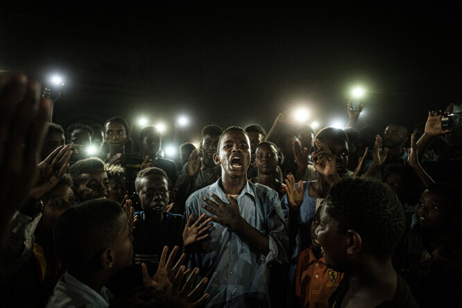 Caption: A young man, illuminated by mobile phones, recites protest poetry while demonstrators chant slogans calling for civilian rule, during a blackout in Khartoum, Sudan, on 19 June 2019.Story: Protests in Sudan began in December 2018 and spread rapidly throughout the country. By April 2019, protesters were staging a sit-in close to army headquarters in the capital Khartoum, and demanding an end to the 30-year rule of dictator Omar al-Bashir. On 11 April, al-Bashir was removed from office in a military coup, and a transitional military government was established. Protests continued, calling for power to be handed to civilian groups. On 3 June, government forces opened fire on unarmed protesters. Scores of people were killed and many more subject to further violence. Three days later the African Union suspended Sudan, in the midst of widespread international condemnation of the attack. The authorities sought to defuse protests by imposing blackouts, and shutting down the internet. Protesters communicated by text message, word of mouth and using megaphones, and resistance to military rule continued. Despite another severe crackdown on 30 June, the pro-democracy movement was eventually successful in signing a power-sharing agreement with the military, on 17 August.